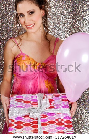 Young woman holding a wrapped gift and a pink balloon against a silver glitter background at a birthday party, smiling.