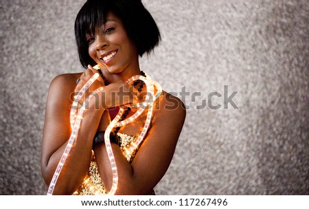 Close up portrait of a beautiful black woman holding Christmas fairy lights around her neck and smiling against a silver glitter background.