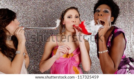 Three young women having a party and blowing whistles on each other, against a silver background.