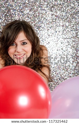 Portrait of an attractive young woman against a silver glitter background, holding two balloons in red and pink colors, smiling.