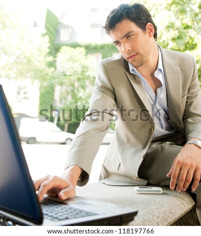 Attractive young businessman using a laptop computer while sitting on a bench in the financial district of a classic city, working outdoors.