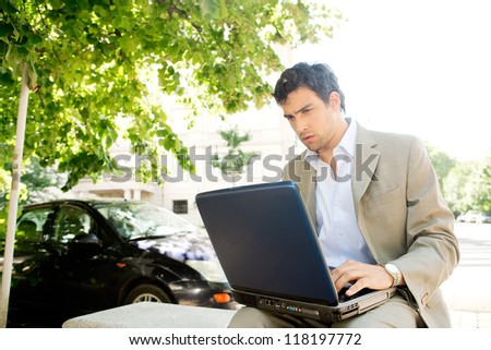 Attractive young businessman using a laptop computer while sitting on a bench in the financial district of a classic city, working outdoors.