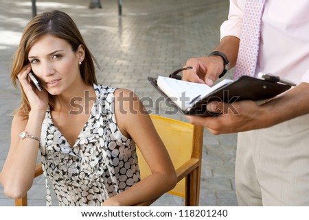 Young businesswoman making a phone call on her cell while a colleague businessman is holding a filofax with notes next to her, outdoors.