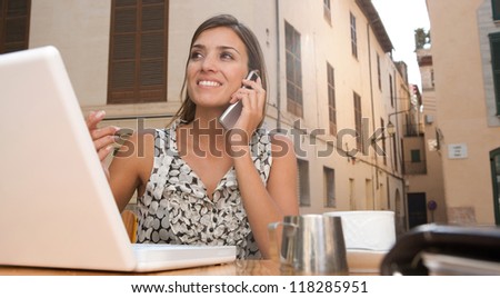 Attractive young businesswoman using a cell phone to make a call while using a laptop computer, sitting at a coffee shop terrace, outdoors.