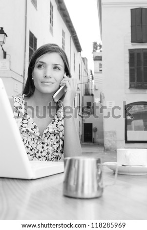 Black and white portrait of an attractive young businesswoman using a cell phone to make a call while using a laptop computer, sitting at a coffee shop terrace outdoors, smiling.