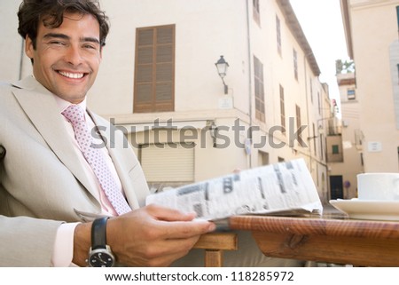 Attractive businessman reading the newspaper in a coffee shop terrace table in a classic city, smiling at the camera, outdoors.
