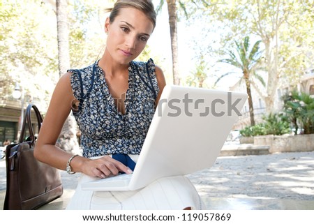 Young businesswoman using a laptop computer while sitting on a bench in a city with palm trees, outdoors.