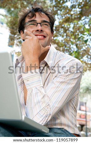 Close up portrait of a young businessman using a laptop computer while sitting on a bench in a city park, outdoors.
