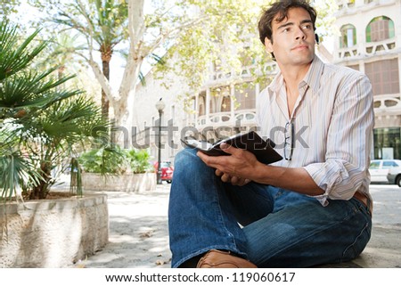Young attractive businessman using a hands free device to make a call on his cell phone while sitting on a bench in a city square with classic architecture.