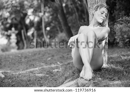 Black and white view of a beautiful nude woman in a nature environment, leaning on a tree trunk and relaxing on grass.