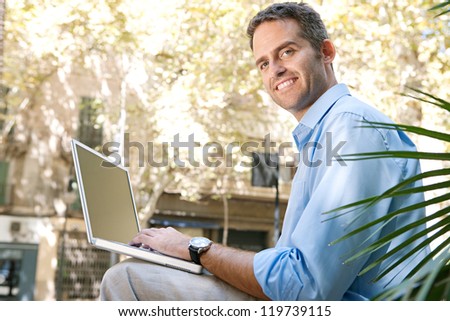 Senior businessman using a laptop computer while sitting on a bench in the city, turning to camera.