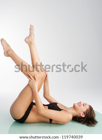 Side view of a young woman laying down on a reflective surface with her legs up in the air, wearing a black swimming costume and smiling.