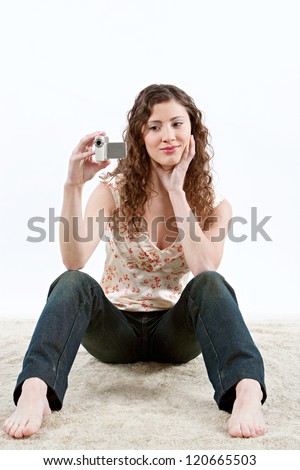 Teenage girl using a small video camera to film while sitting down on a furry carpet at home, smiling.
