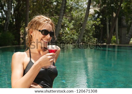 Fashionable young woman drinking a cocktail while sitting by the edge of a tropical swimming pool wearing a black swimming costume.