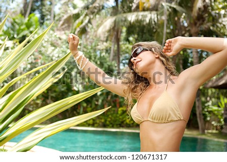 Beautiful young woman dancing in a golden bikini near a swimming pool in a topical garden during a sunny day on vacations.