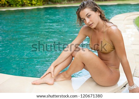 Sexy young woman sitting on the edge of a swimming pool, wearing a gold bikini while on vacations in a sunny destination.