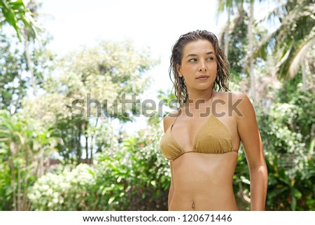 Feminine young woman wearing a golden bikini and standing in a lush forest garden while on vacation in a tropical destination.