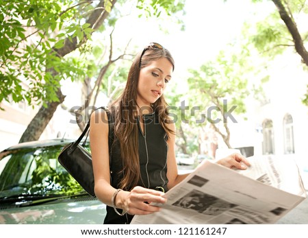 Beautiful young businesswoman reading a newspaper while leaning on a car in the financial district of a classic city street aligned with trees on a sunny day.