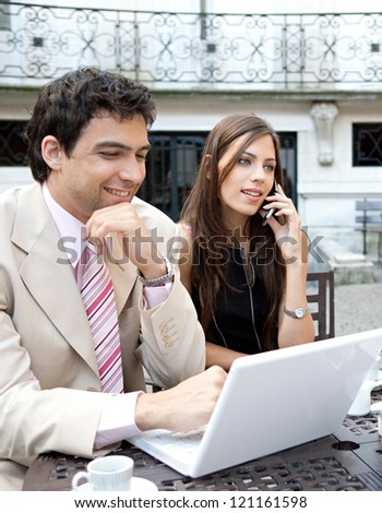 Portrait of two busy business people having a meeting in a coffee shop terrace in a classic city financial district, using technology and smiling.