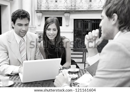 Black and white portrait of three business people sharing a table at a coffee shop terrace, having a meeting and talking while using technology in the financial city district.