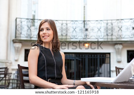 Attractive young businesswoman sitting with her laptop at a coffee shop near classic office buildings in the city, smiling.