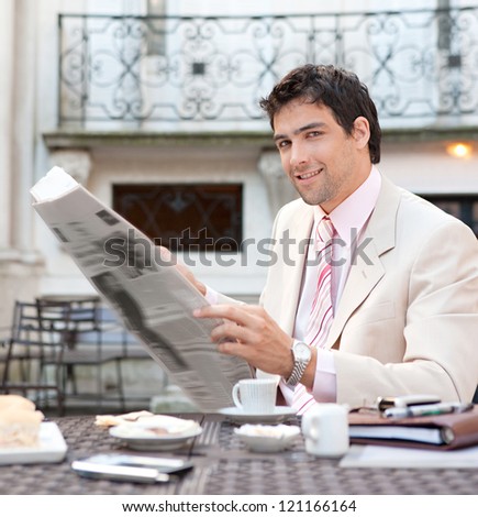 Young attractive hispanic businessman reading the newspaper while having breakfast at a coffee shop terrace in a classic city.
