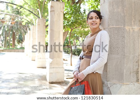 Portrait of an attractive young woman holding her shopping bags while leaning on a stone column in a romantic garden, being thoughtful outdoors.