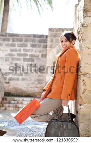 Attractive hispanic young woman leaning on an old stone wall while visiting a sight on vacations, holding shopping bags and smiling.