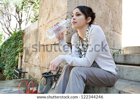 Side view of a young businesswoman sitting on a park steps drinking water from a plastic blue bottle.