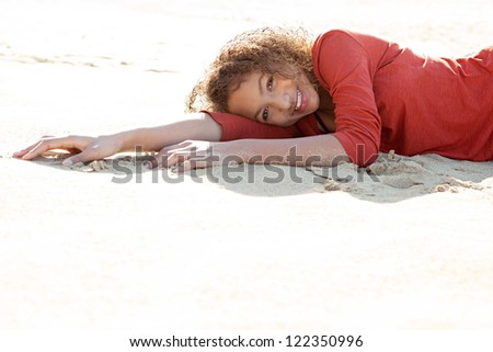 Beauty portrait of an adolescent mixed race girl lying down on a white sand beach with the sun shining on her and smiling at the camera.