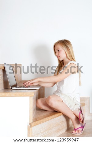 Young child girl using a laptop computer while sitting down on her home\'s wooden stairs and wearing pink flip flop sandals.