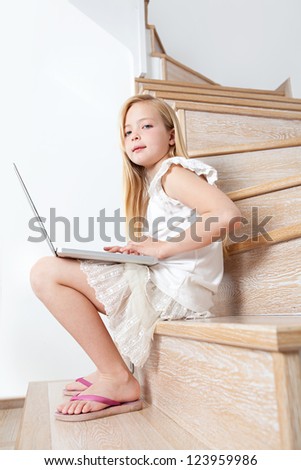 Profile view of a young girl child using a laptop computer while sitting down on her home\'s wooden stairs and wearing pink sandals.