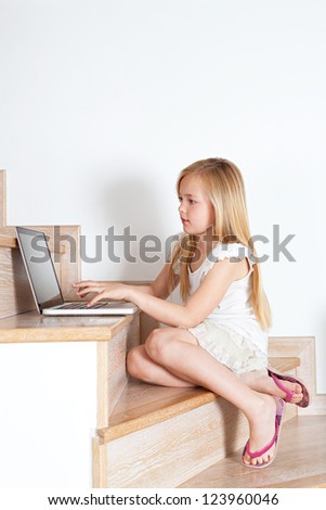 Young child girl using a laptop computer while sitting down on her home\'s wooden stairs and wearing pink flip flop sandals.
