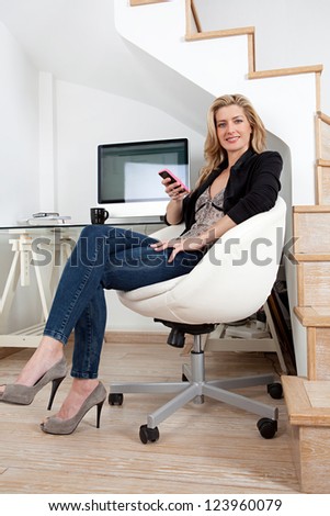 Attractive businesswoman working from home office and using a smart phone while sitting on a modern chair and working at her desk.