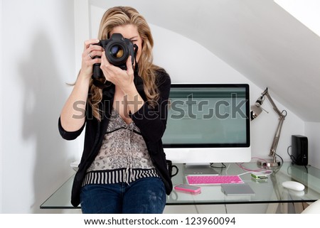 Smart woman photographer taking a photograph with her professional digital photographic camera, focusing the lens while leaning on her work desk in her modern technology office.