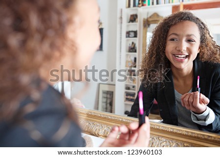Teenage girl applying lipstick using her bedroom mirror and joyfully smiling.