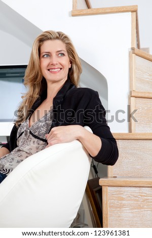Attractive businesswoman sitting down on her home office chair, next to a wood floor stairwell, smiling.