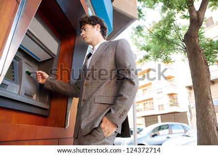Side low view of an elegant businessman withdrawing money from a wood decorated bank cash point, outdoors.