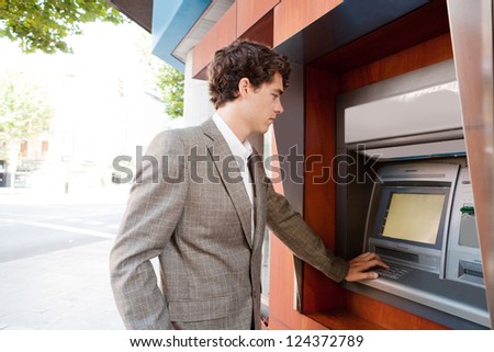 Side view of an elegant businessman withdrawing money from a wood decorated bank cash point in the city, outdoors.