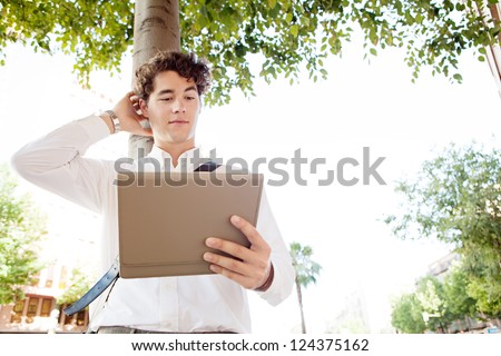 Unsure businessman scratching his head while using a tablet device, standing in a wide city avenue with trees, outdoors.