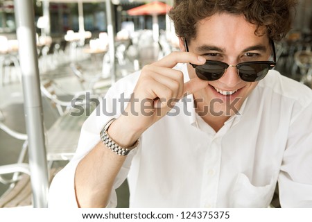 Close up portrait of a young businessman holding his shades to look at the camera smiling, while sitting at a coffee shop terrace, outdoors.