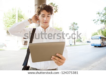 Indecisive businessman scratching his head while using a tablet device, standing in a wide city avenue with traffic and public transport, outdoors.
