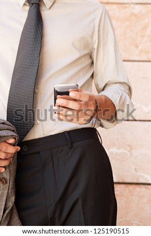 Close up detail of a businessman body section and hand holding a suit jacket and using a smart phone by an office building in the city.