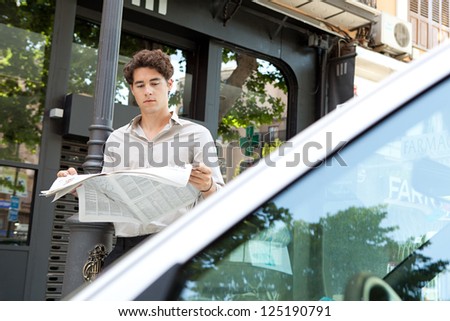 Attractive businessman leaning on a lamp post in the city behind a car, reading the newspaper.