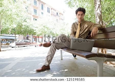Businessman sitting on a wooden bench in the city using his laptop. Low perspective view.