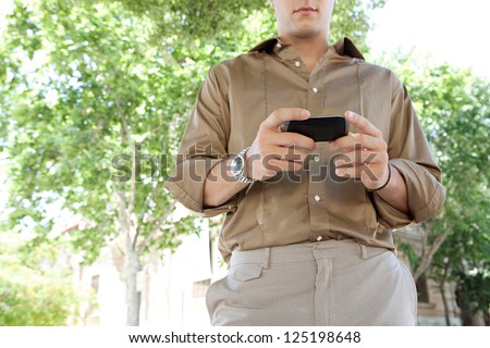 Middle body section of a businessman using a smart phone while standing near office buildings and green trees in the city, outdoors.