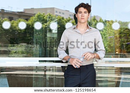 Young businessman leaning on an office building glass window holding a bottle of mineral water, with reflections of the city behind him.