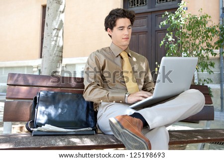 Portrait of a young smart businessman sitting on a wooden bench in the city and using his laptop computer, outdoors.