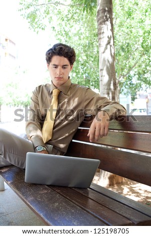 Smart businessman sitting on a wooden bench in a city park and using his laptop computer, outdoors.