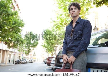 Young relaxed businessman leaning on a car, waiting while holding his leather briefcase and wearing elegant clothes.